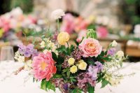 a colorful wedding centerpiece of pink peonies, pink and yellow ranunculus, lilac blooms and greenery, herbs and candles around