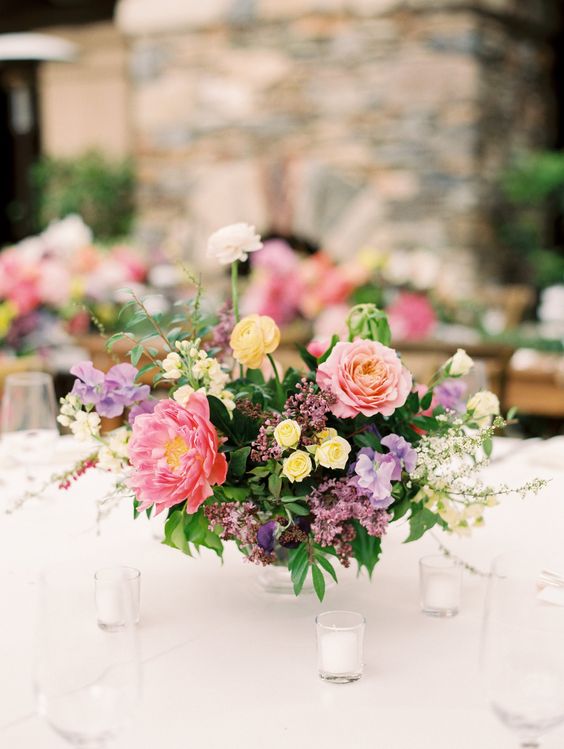 a colorful wedding centerpiece of pink peonies, pink and yellow ranunculus, lilac blooms and greenery, herbs and candles around