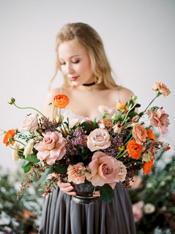 a dimensional and colorful wedding centerpiece of mauve and blush roses, orange ranunculus, greenery and berries and some lilac fillers