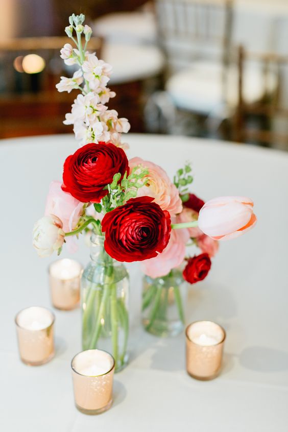 a lovely and pretty wedding centerpiece of clear vases, pink and red ranunculus and peachy tulips plus candles is a very stylish and bold solution