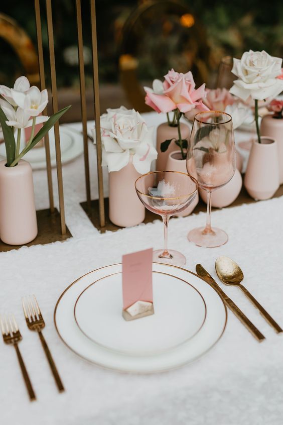 a lovely modern wedding tablescape with a neutral tablecloth, blush vases with a single bloom in each, gold rimmed plates and gold cutlery