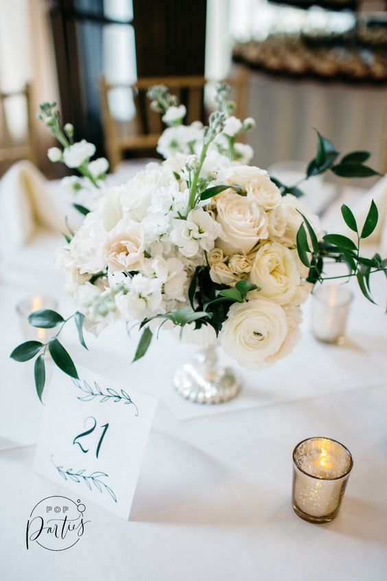 a lush white floral centerpiece of ranunculus, roses and other blooms, greenery plus a refined silver bowl and lots of candles around