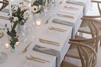 a modern and laconic wedding table setting with neutral linens, white blooms and greenery, tall and thin candles, smaller candles and gold cutlery