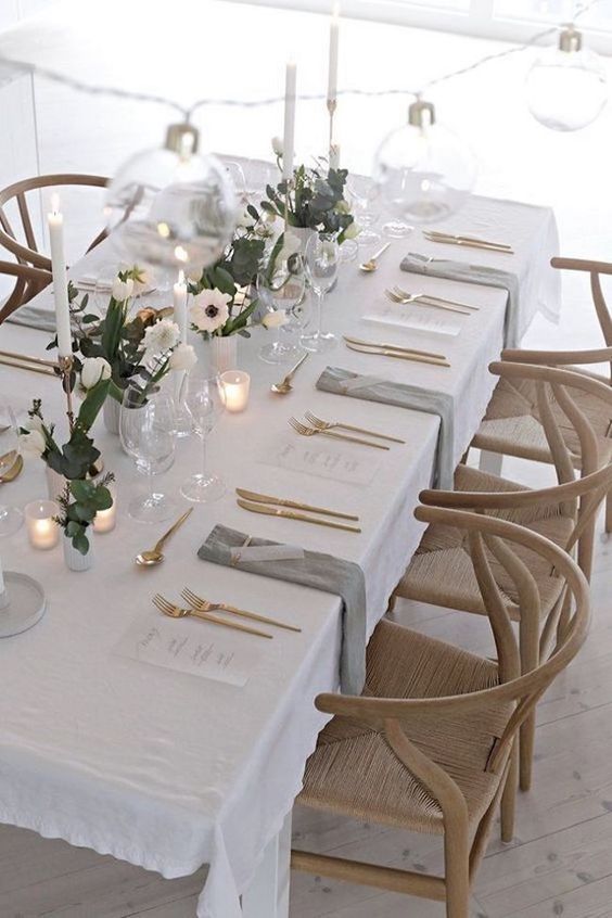 a modern and laconic wedding table setting with neutral linens, white blooms and greenery, tall and thin candles, smaller candles and gold cutlery