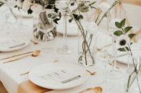 a modern neutral wedding tablescape with a white tablecloth, tan napkins, white porcelain, gold cutlery and white blooms and greenery