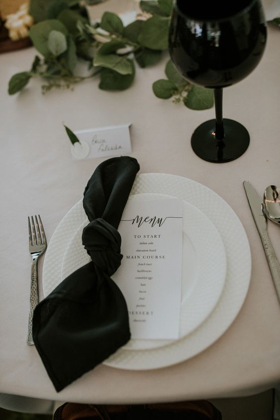 a pretty modern wedding table setting with a white tablecloth and a black napkin, a black glass and a greenery runner, white porcelain