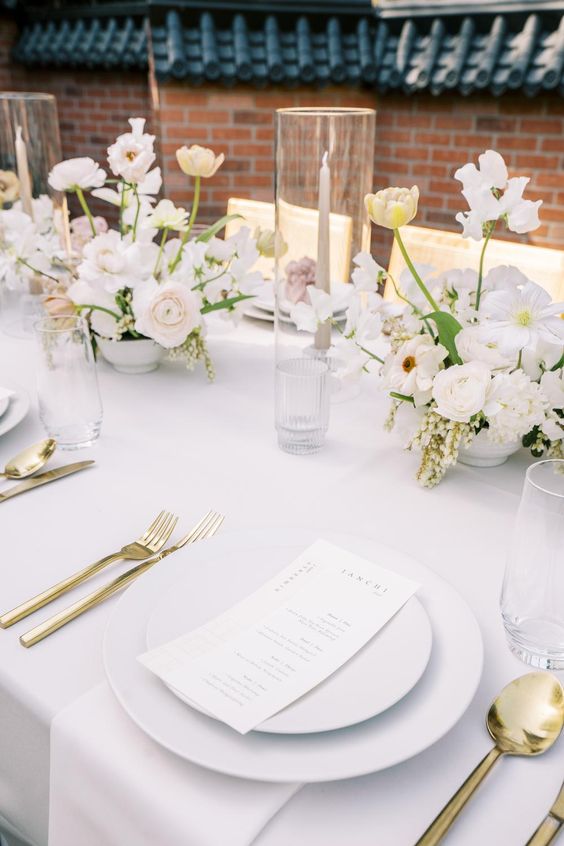 a refined modern wedding table setting with white linens and porcelain, a blush, white and yellow floral centerpiece and gold cutlery