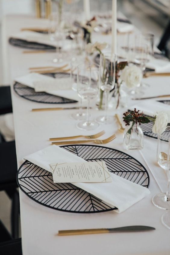 a refined modern wedding tablescape with geometric black placemats, gold cutlery, white blooms and leaves and tall and thin candles