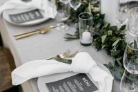 a stylish modern wedding tablescape with a grey tablecloth and white napkins, a greenery runner with pillar candles and gold cutlery