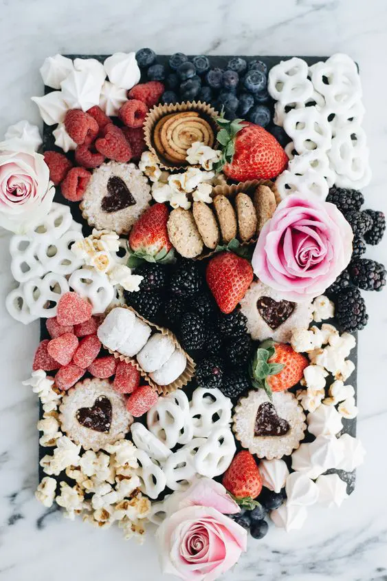 a sophisticated wedding grazing table with glazed crackers, popcorn, blackberries, bluberries, raspberries, cookies and pink roses is a beautiful idea