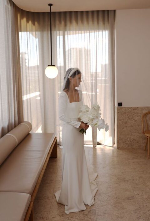 a stylish minimalist bridal look with a square neckline mermaid wedding dress, long sleeves and a train, a pearl headpiece and a veil