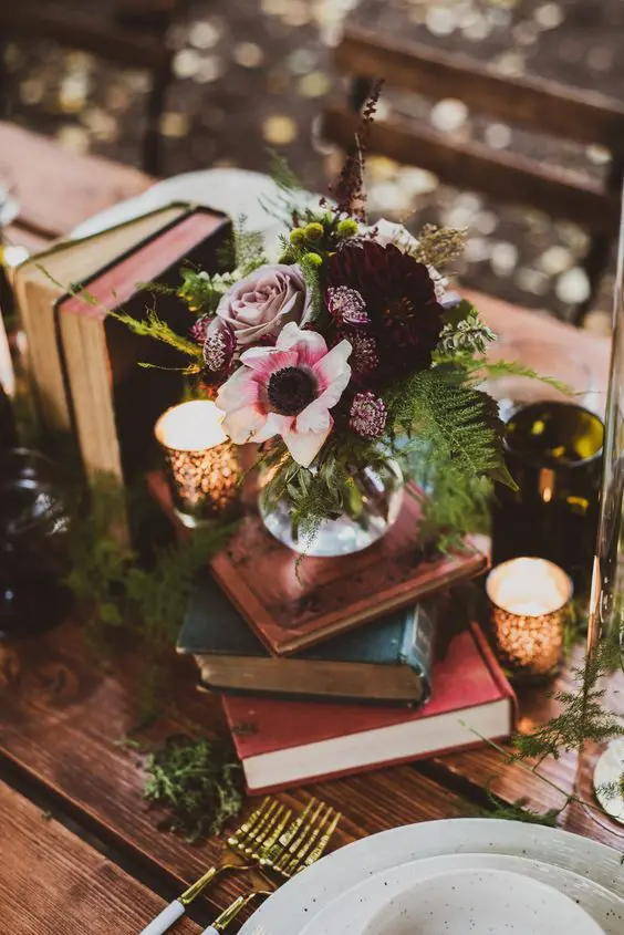 a beautiful dark enchanted forest wedding centerpiece of a stack of books, candles, a dark floral centerpiece and greenery