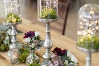 a cool enchanted forest wedding centerpiece of moss, tall closhes with moss and lights, white and burgundy blooms in vases
