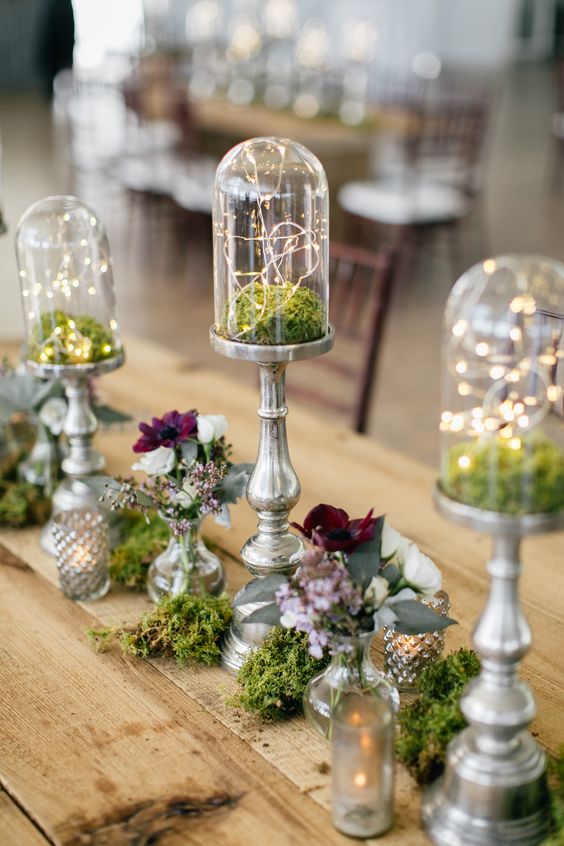 a cool enchanted forest wedding centerpiece of moss, tall closhes with moss and lights, white and burgundy blooms in vases