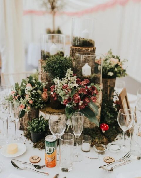 a creative enchanted forest wedding centerpiece composed of tree sutmps, burgundy and pastel blooms and greenery, moss and candles