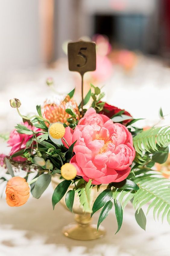 a gorgeous bright summer wedding centerpiece of a pink peony, billy balls, orange roses, a pincushion protea and a table number is adorable