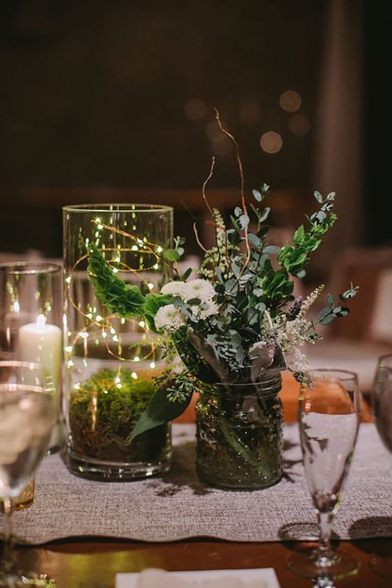 a lovely cluster wedding centerpiece of a jar with greenery and white blooms, a jar with moss and lights and candles around