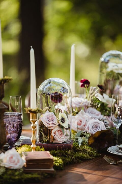 a lovely enchanted forest wedding centerpiece of moss, blush blooms, vintage candleholders with thin and tall candles and a stack of books