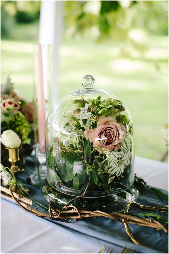 a lovely wedding centerpiece of a cloche filled with greenery and mauve blooms and vines around is a cool enchanted forest wedding centerpiece