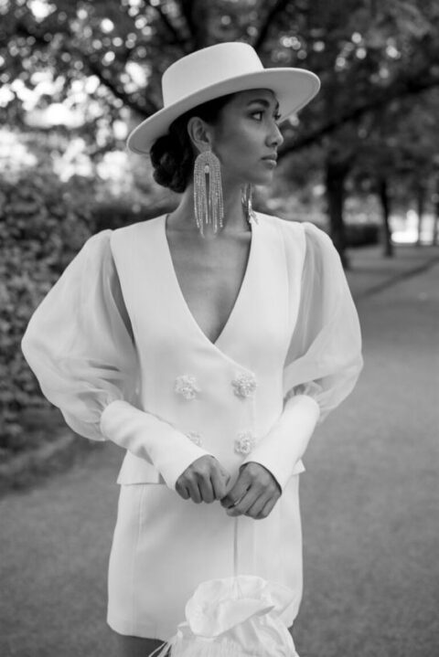 a sophisticated and modern bridal look with a white blazer dress with semi sheer puff sleeves and embellished buttons, statement earrings and a hat