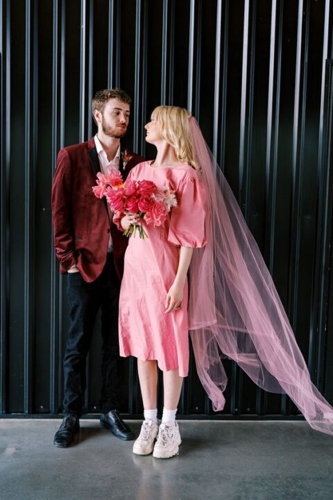 an 80s bridal look with a plain pink midi dress with puff sleeves, white trainers and socks, a pink veil and a pink wedding bouquet