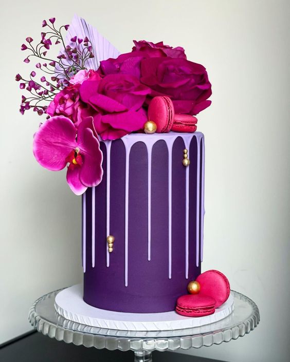a purple wedding cake with lilac drip, gold beads, hot pink and fuchsia blooms and macarons on top, a palm leaf and a bit of blooming branches