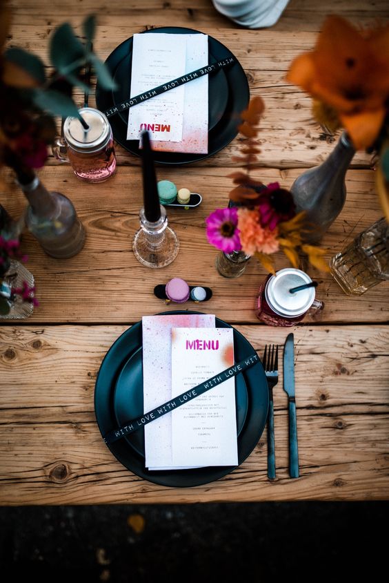 a catchy and bold wedding tablescape in rock n roll style, with an uncovered table, bold blooms and macarons, black plates and colorful menus