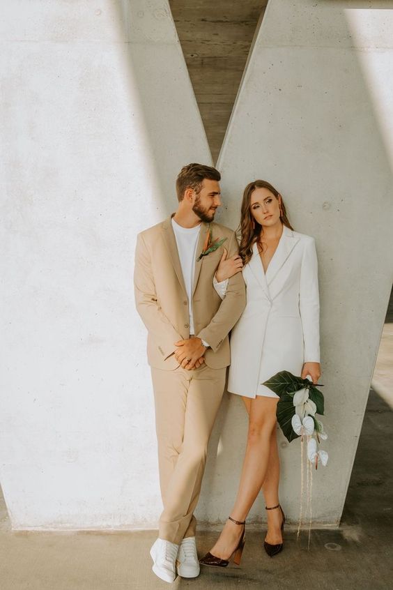 chic minimalist couple's looks with a beige suit with a white t-shirt and white sneakers, a white blazer wedding dress and black shoes