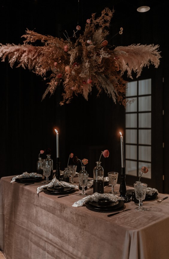 a chic moody rock wedding tablescape with a dusty pink tablecloth, black plates, black vases and candleholders, a grass and bloom overhead isntallation