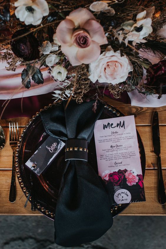 a refined glam rock wedding tablescape with a blush runner, blush and white and deep purple blooms, black chargers and napkins