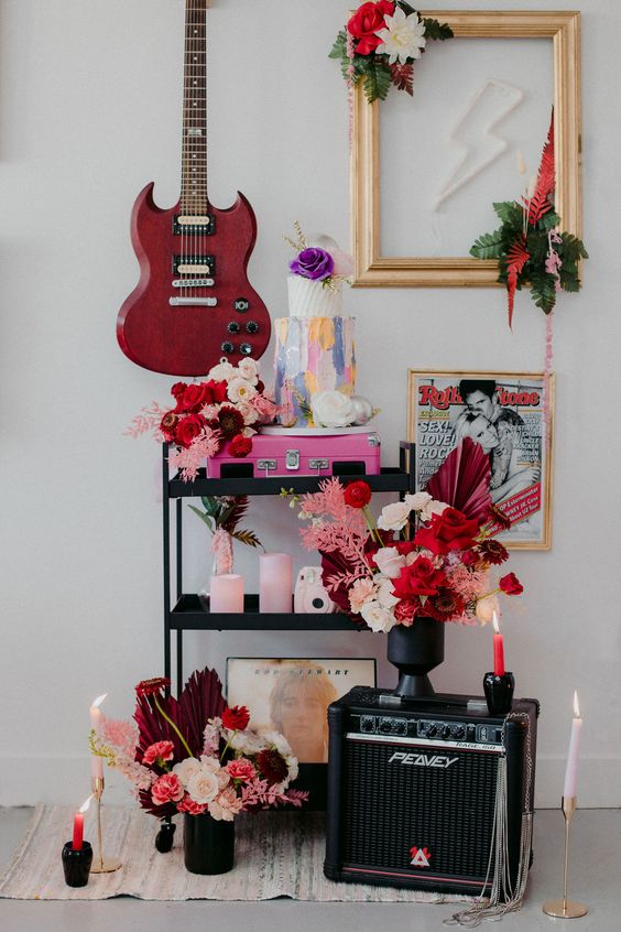 bold rock inspired wedding decor wiht a pink guitar, pink candles, blush and hot red blooms, a colorful cake and a neon sign is wow