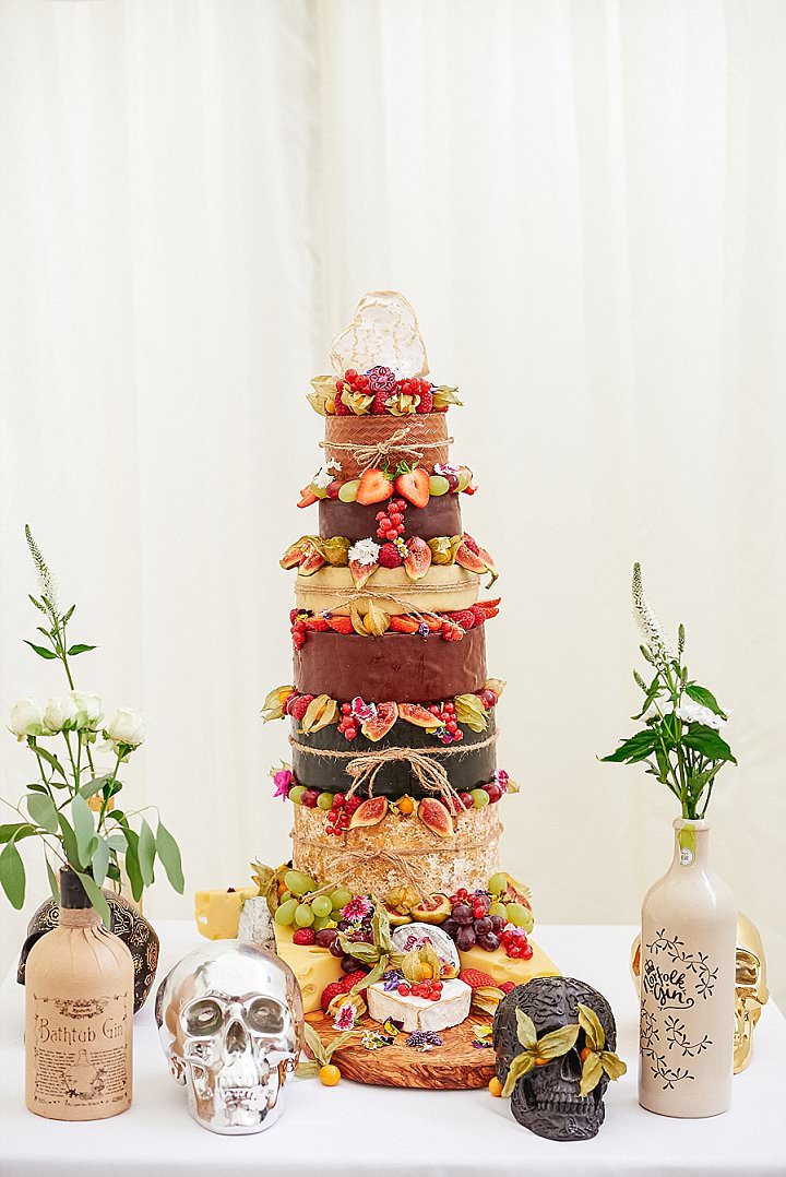 a delicious looking cheese wheel wedding cake decorated with various berries and fruit, surrounded with greenery and skulls