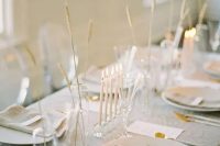 26 a delicate minimalist wedding tablescape with a grey tablecloth and napkins, white chargers and cutlery, some grasses in vases and tall and thin candles