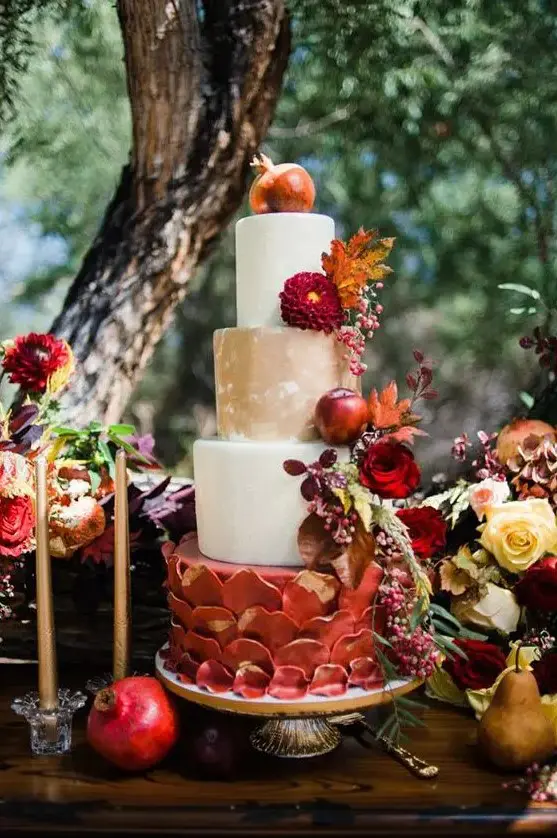 a whimsical wedding cake with white, tan, burgundy petal with gold tiers, with burgundy blooms, berries, fall elaves and apples and a pomegranate on top for a fall wedding