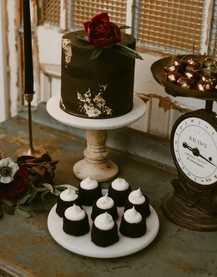 a gorgeous rock n roll dessert table with a black wedding cake with gold leaf, a burgundy bloom, gilded cherries and brownies