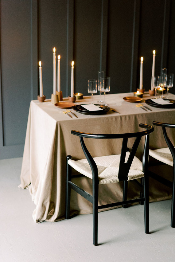 a minimalist fall wedding table setting with a beige tablecloth, terracotta candleholders, black plates and gold cutlery