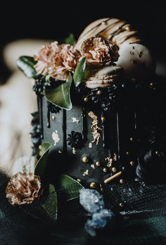 a refined black wedding cake with black and gold beads, black meringues, neutral gilded macarons and blooms plus greenery on top