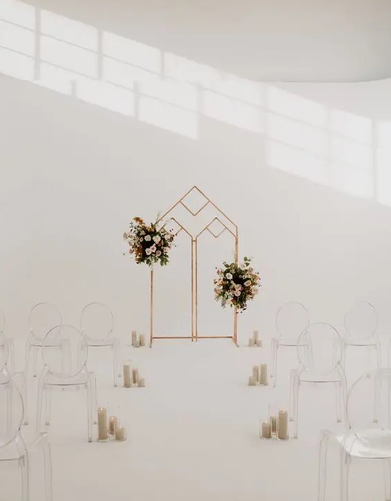 a minimalist wedding ceremony space with a geo wedding arch with pink floral arrangements, ghost chairs and pillar candles lining up the aisle