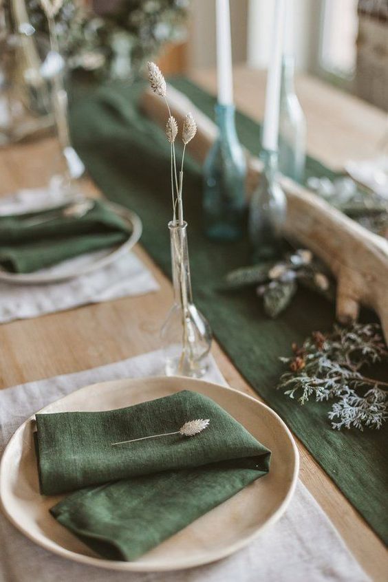 a natural woodland fall wedding tablescape with a green runner and napkins, neutral plates and candles plus some evergreens