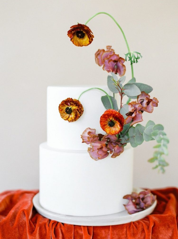 a beautiful wedding cake in white decorated with bodl orange and mauve blooms and greenery is a lovely idea for a modern or minimal fall wedding