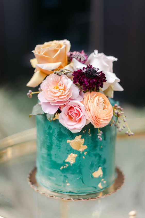 an emerald textural wedding cake with gold foil, with orange, blush and white blooms and purple ones for a bolder touch
