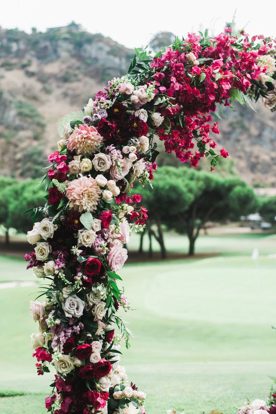 a bold jewel tone wedding arch with deep red, burgundy and fuchsia blooms, white and blush ones plus some greenery