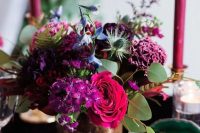 a bold jewel tone wedding centerpiece of purple, pink, deep purple and blue blooms, greenery and thistles and fuchsia candles around