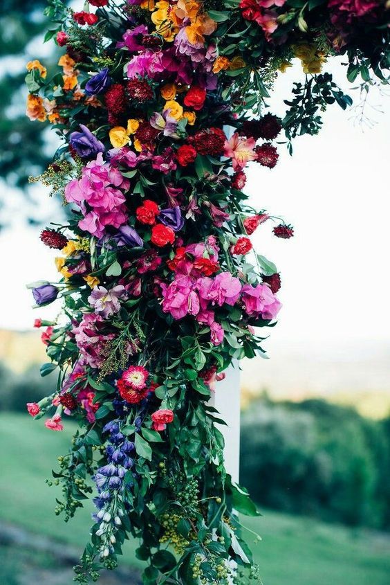 a bold wedding arch decorated with purple, pink, red, burgundy and yellow blooms and greenery for a jewel tone wedding
