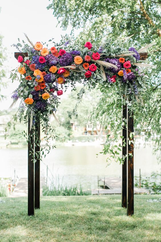 a bright wedding arch featuring purple, yellow, orange and hot pink blooms, greenery and pampas grass for a bold fall wedding