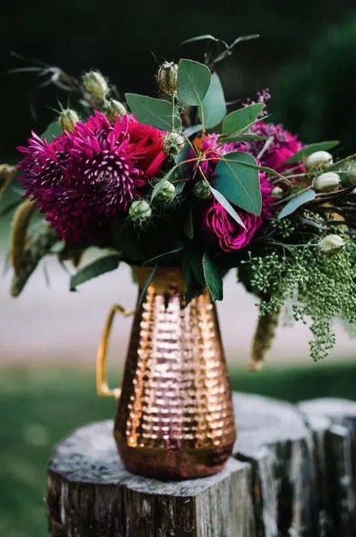 a chic fall wedding centerpiece of a copper pitcher, lush greenery and plum-colored blooms for a wow look