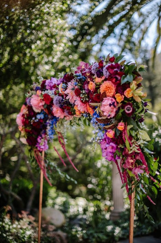 a colorful wedding arch in jewel tones - burgundy, deep red, orange, yellow and pink blooms and greenery is a chic idea for a fall wedding