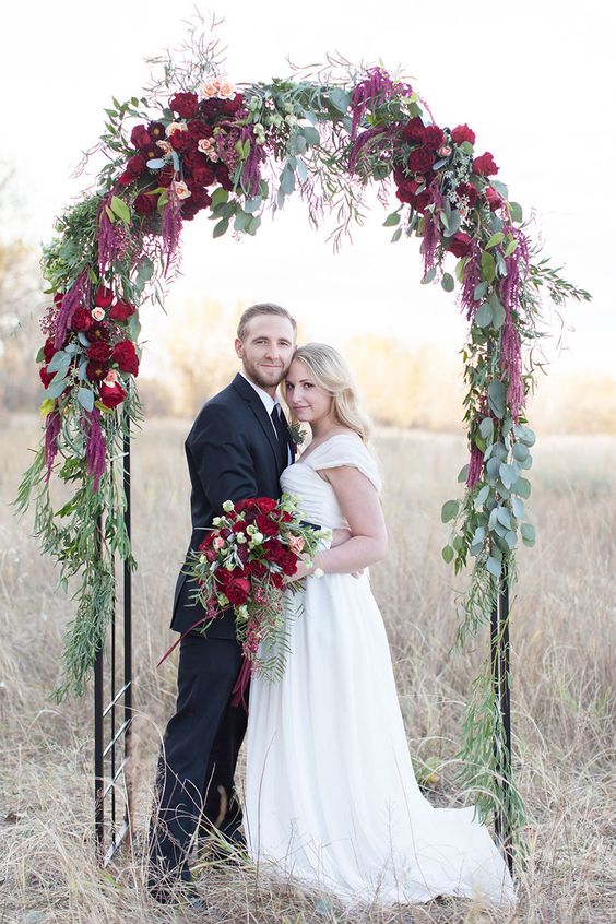 a delicate fall wedding arch decorated with peachy blooms, burgundy ones, deep red blooms and lots of greenery