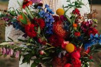 a fantastic jewel tone wedding bouquet with red, orange, blue, lilac blooms, greenery, billy balls and much texture