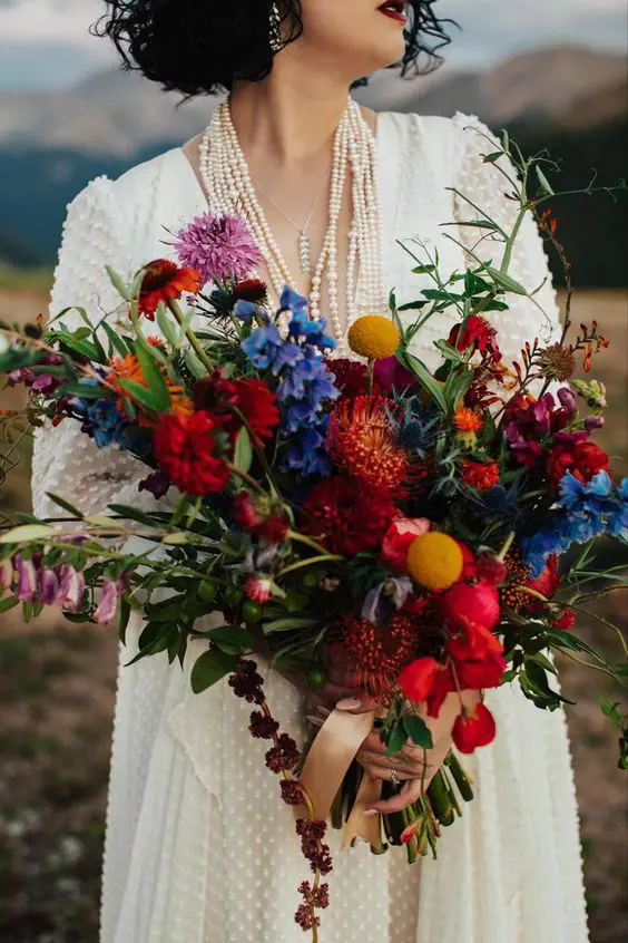 a fantastic jewel tone wedding bouquet with red, orange, blue, lilac blooms, greenery, billy balls and much texture